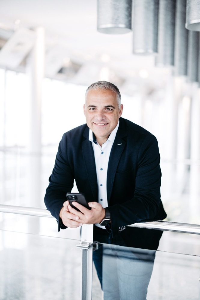Enrico Ender, Gründer von Praevisio Digital, lehnt lächelnd an einem Glasgeländer und hält ein Smartphone.